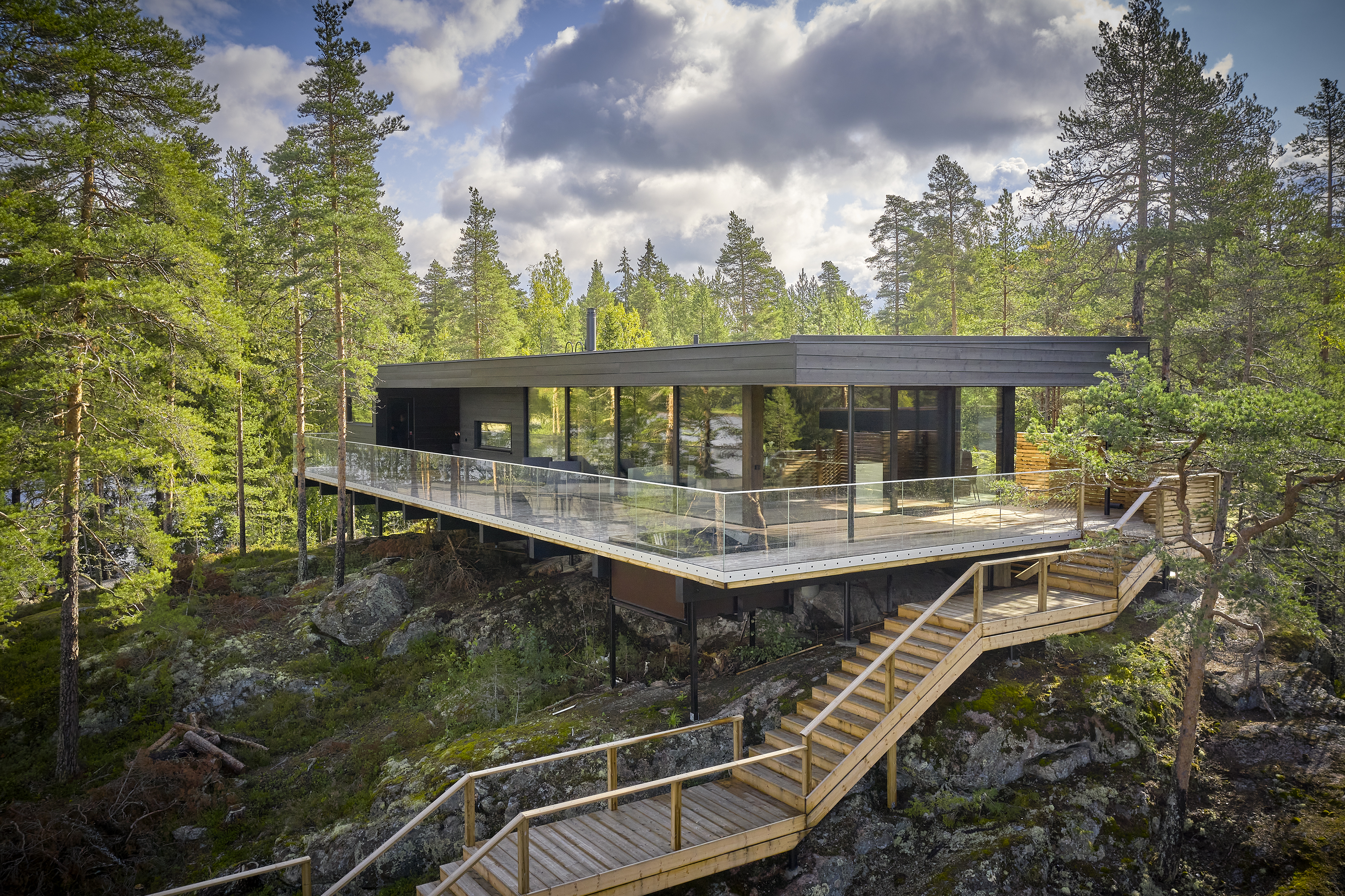 Black timber villa with a wide glass-railed terrace set into a rocky pine forest slope